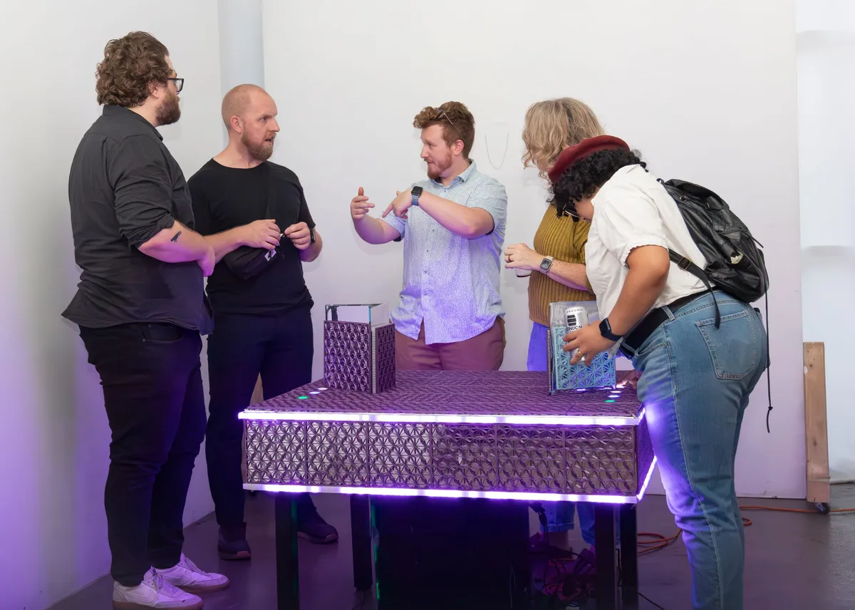 Visitors standing around a light-edged plinth holding one of the installations, drinks in hand, mid-conversation.