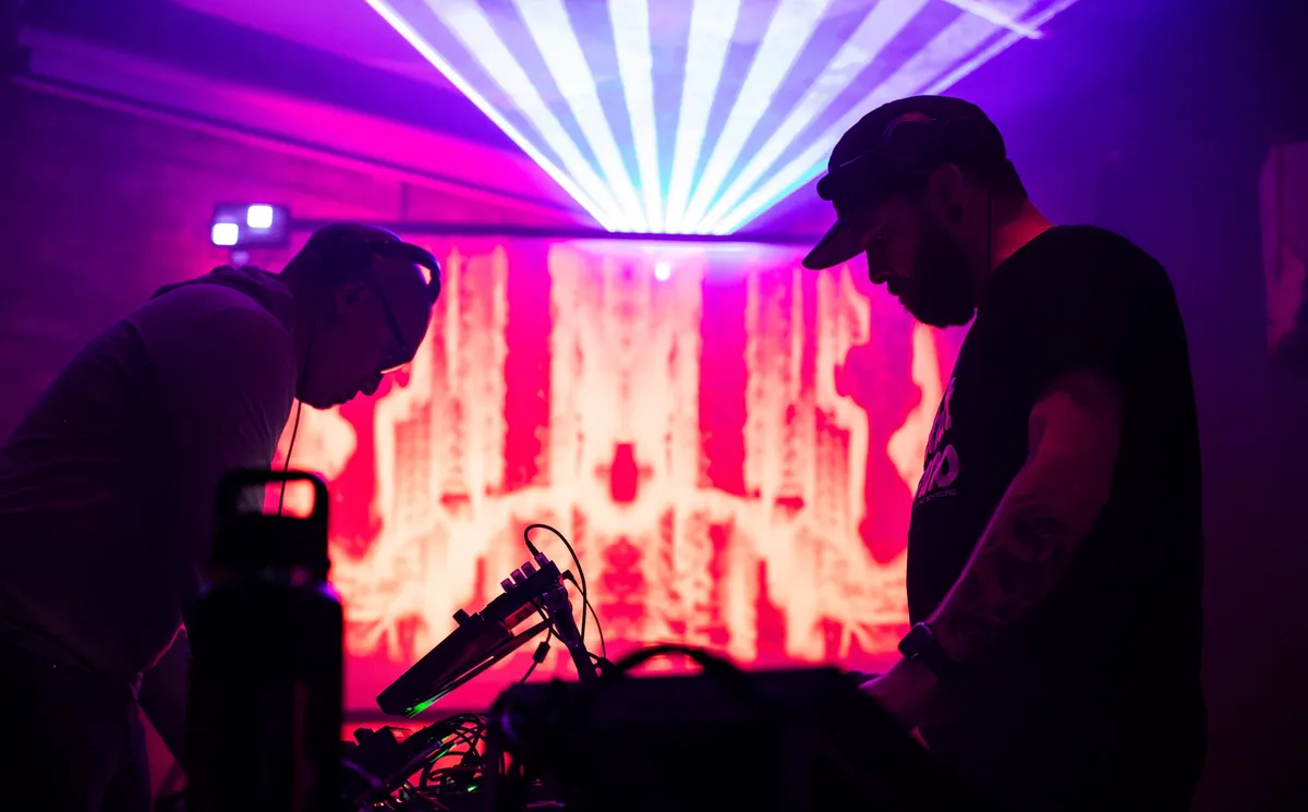 Two artists bent over their gear at A/V Nights 2, red and white visuals filling the wall behind them and a fan of magenta and cyan light thrown across the ceiling.