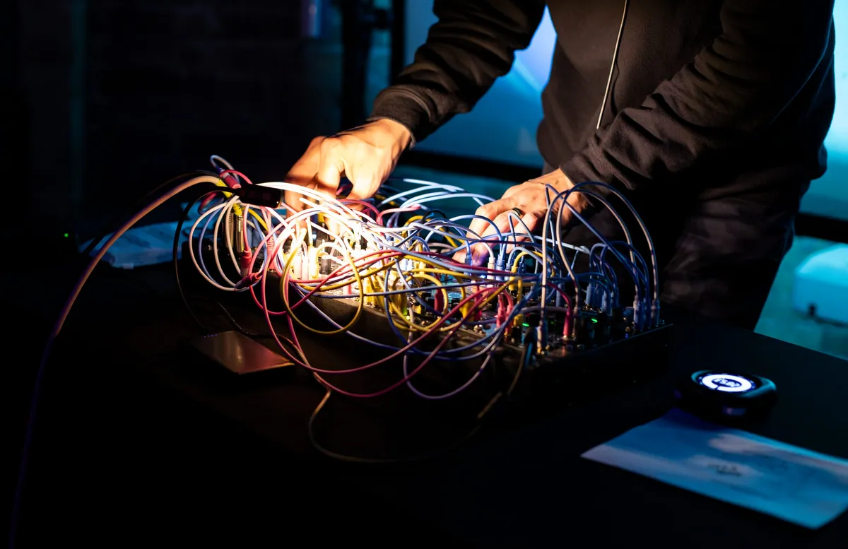 Hands on a modular synthesizer, a dense tangle of patch cables lit from within on a dark table.