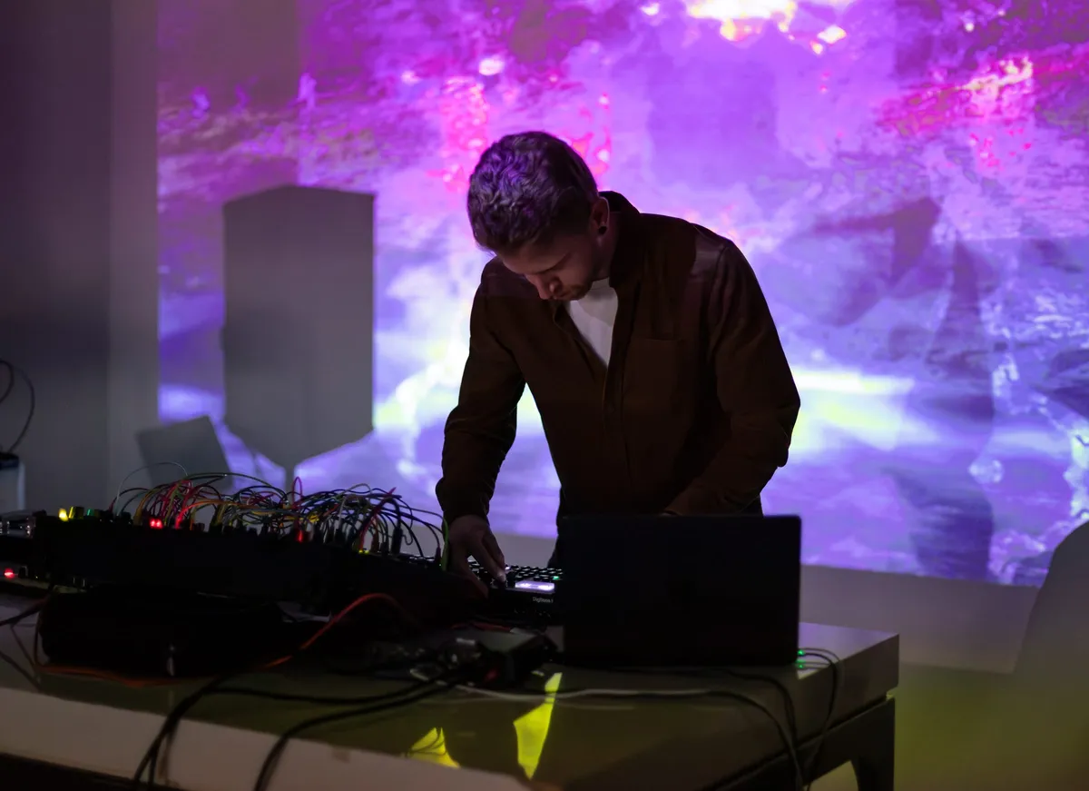 A performer working a bank of synthesizers in front of a magenta and violet projection that fills the wall behind them.