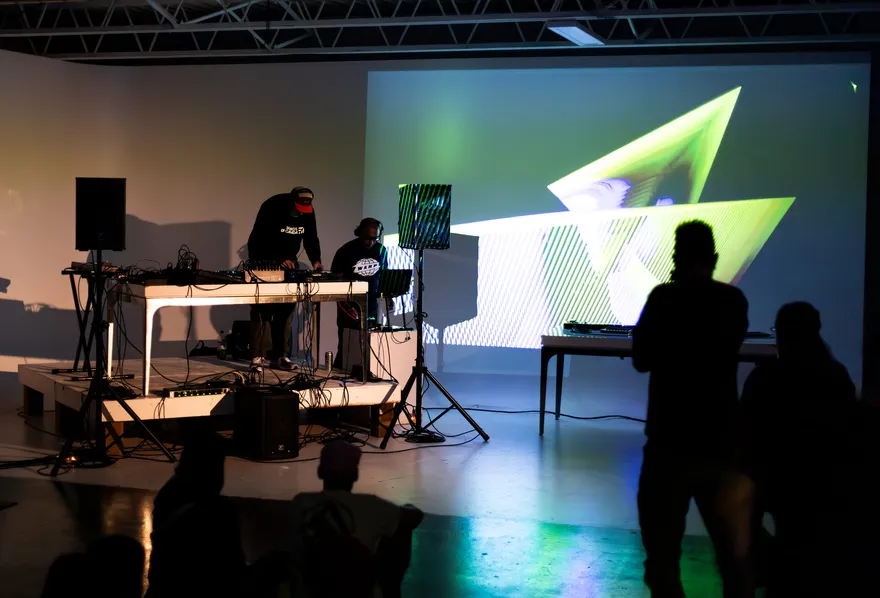 A darkened studio at A/V Nights 1 — two performers at a table of gear beside a wall-sized green geometric projection, the audience watching in silhouette.