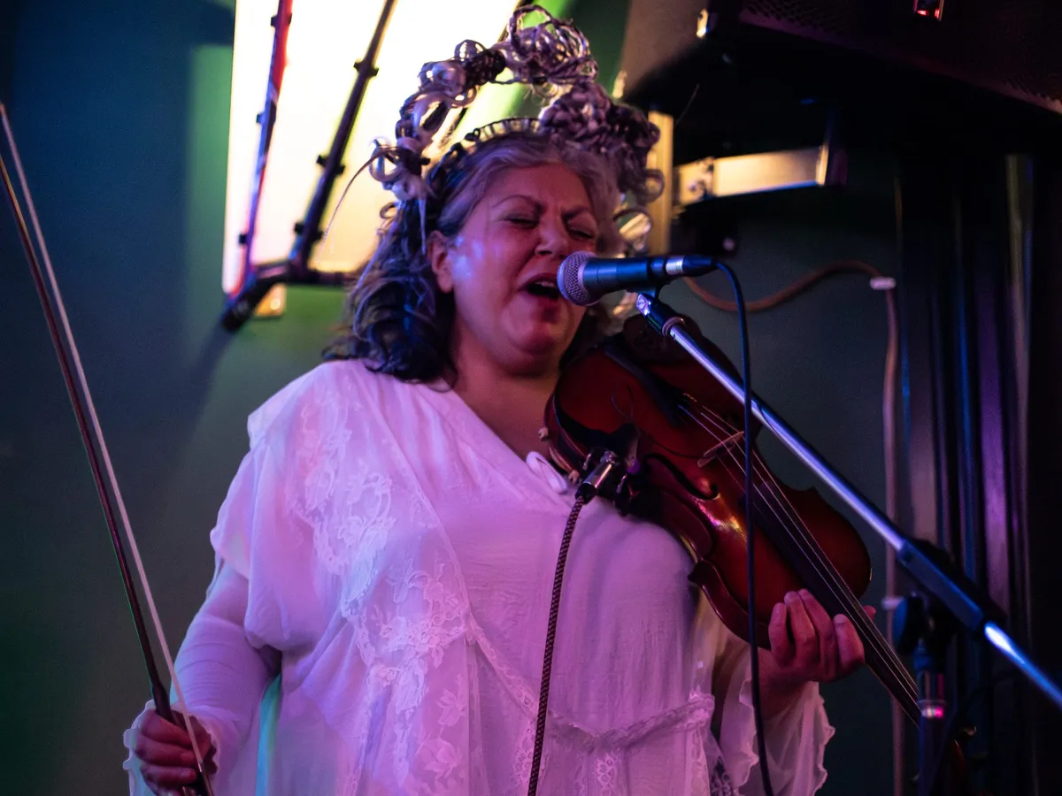 A performer in a white dress and a wire flower crown singing into a microphone while playing violin under stage light.