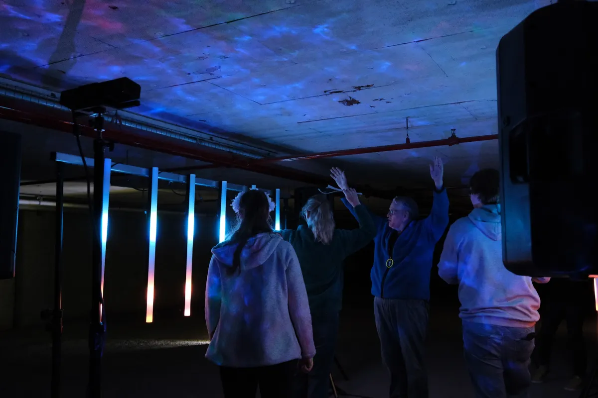 Seen from behind, four visitors raising their arms toward a row of glowing tubes, with rippling blue light spread across the concrete ceiling above.
