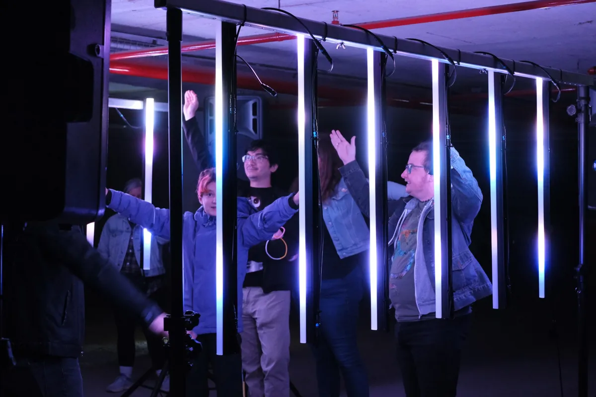A row of hanging white light tubes in front of four visitors, arms stretched out and raised, everyone looking up at the light.