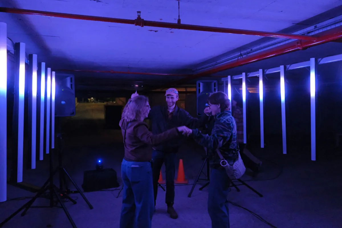 Three people holding hands and turning in a circle in blue light between two rows of white columns, an orange traffic cone behind them.