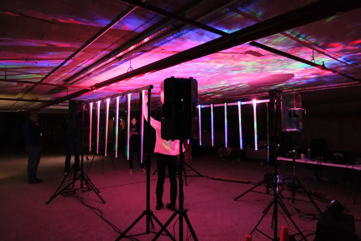 The whole installation from outside — two frames of light tubes, speakers on tripods, and the garage ceiling washed in magenta and green.