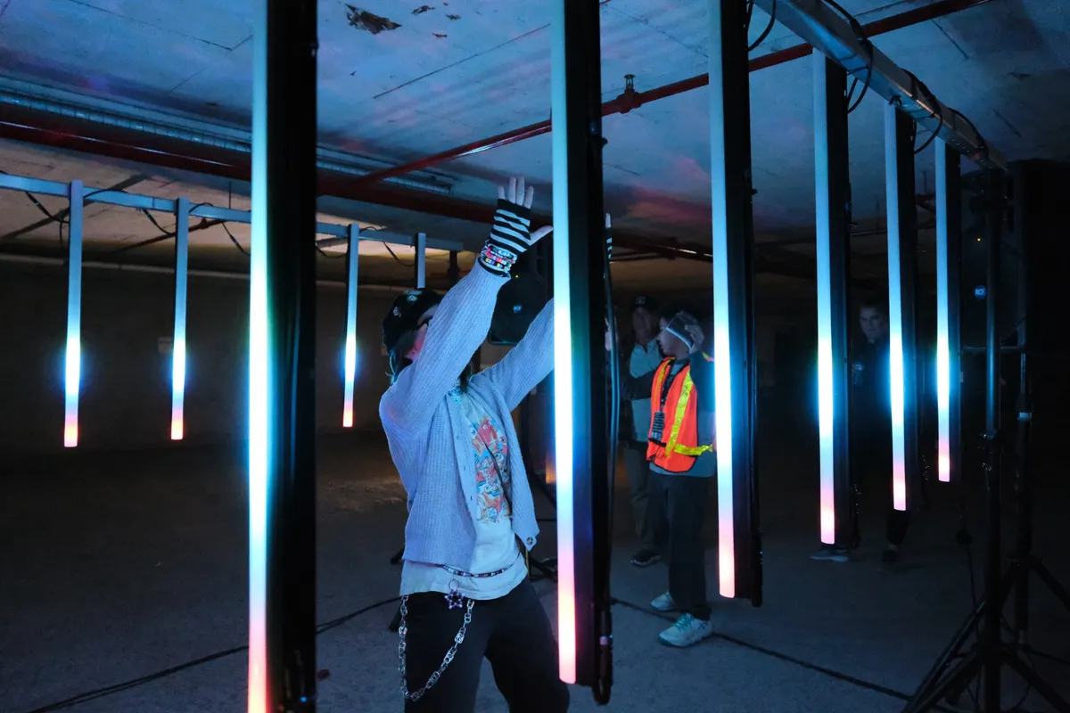 A visitor in a striped glove and beaded bracelets reaching up between tall tubes that fade from cyan to pink at their tips.