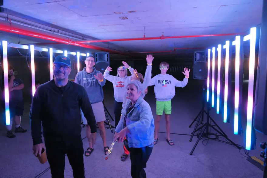 Visitors with their hands raised between two rows of glowing LED columns in the underground garage, lit in cyan, white and magenta, speakers on stands at either side.
