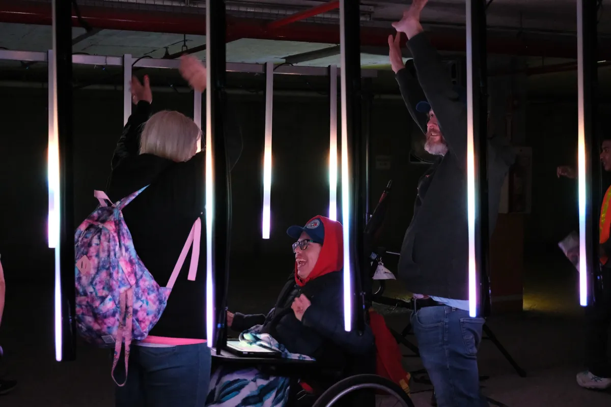 A visitor in a wheelchair, in a red hood and ball cap, laughing up at the light while two people beside them throw their arms overhead among the glowing tubes.