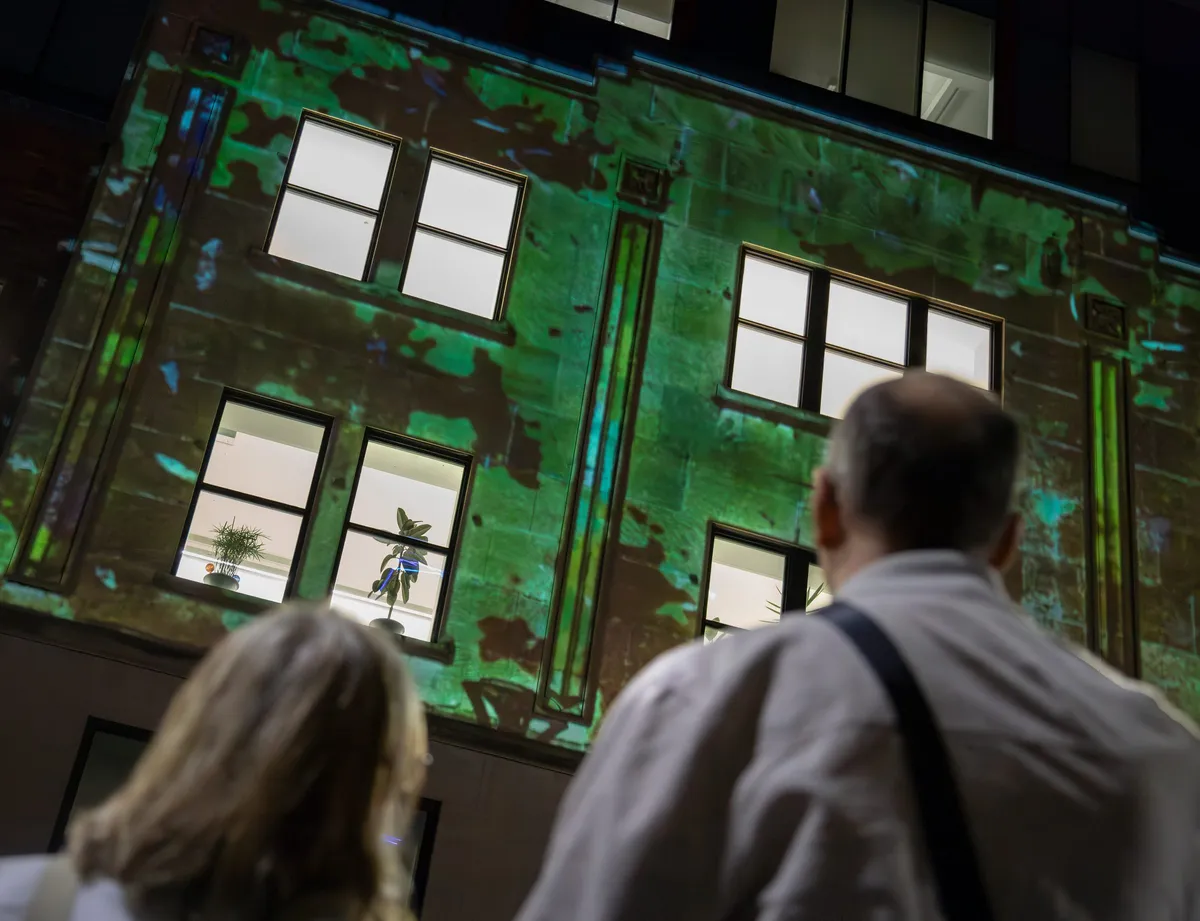 Green foliage spreading across the lit stone of the façade while people watch from below.