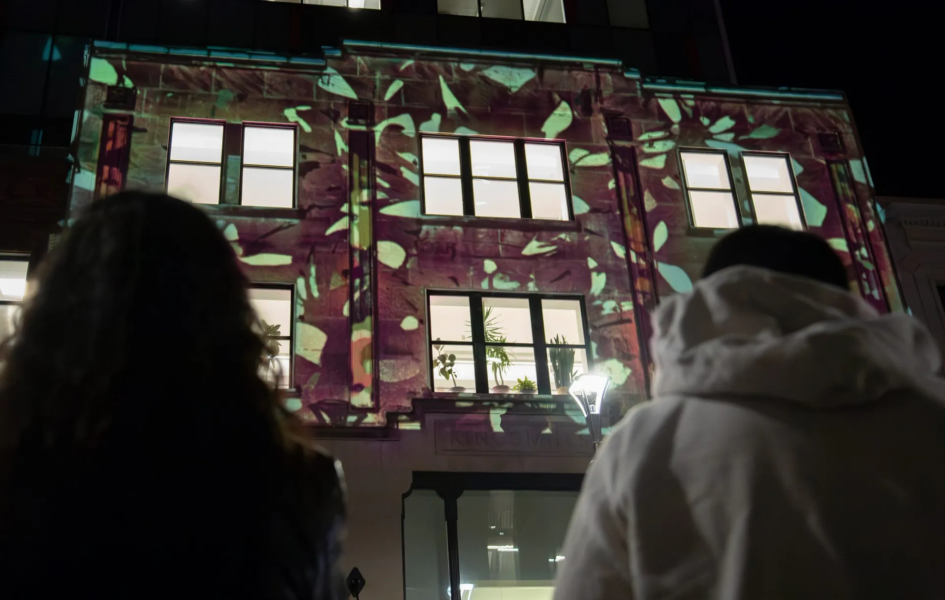 The Kingsmill's façade at night, three storeys washed in magenta and green foliage, with two people watching from the street below.