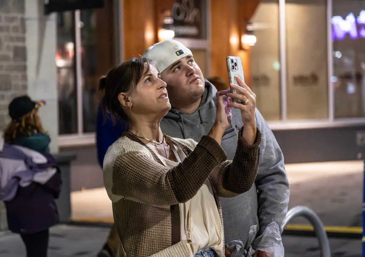 Two visitors on the sidewalk holding up a phone, looking up at the projection as they take their turn at the controls.