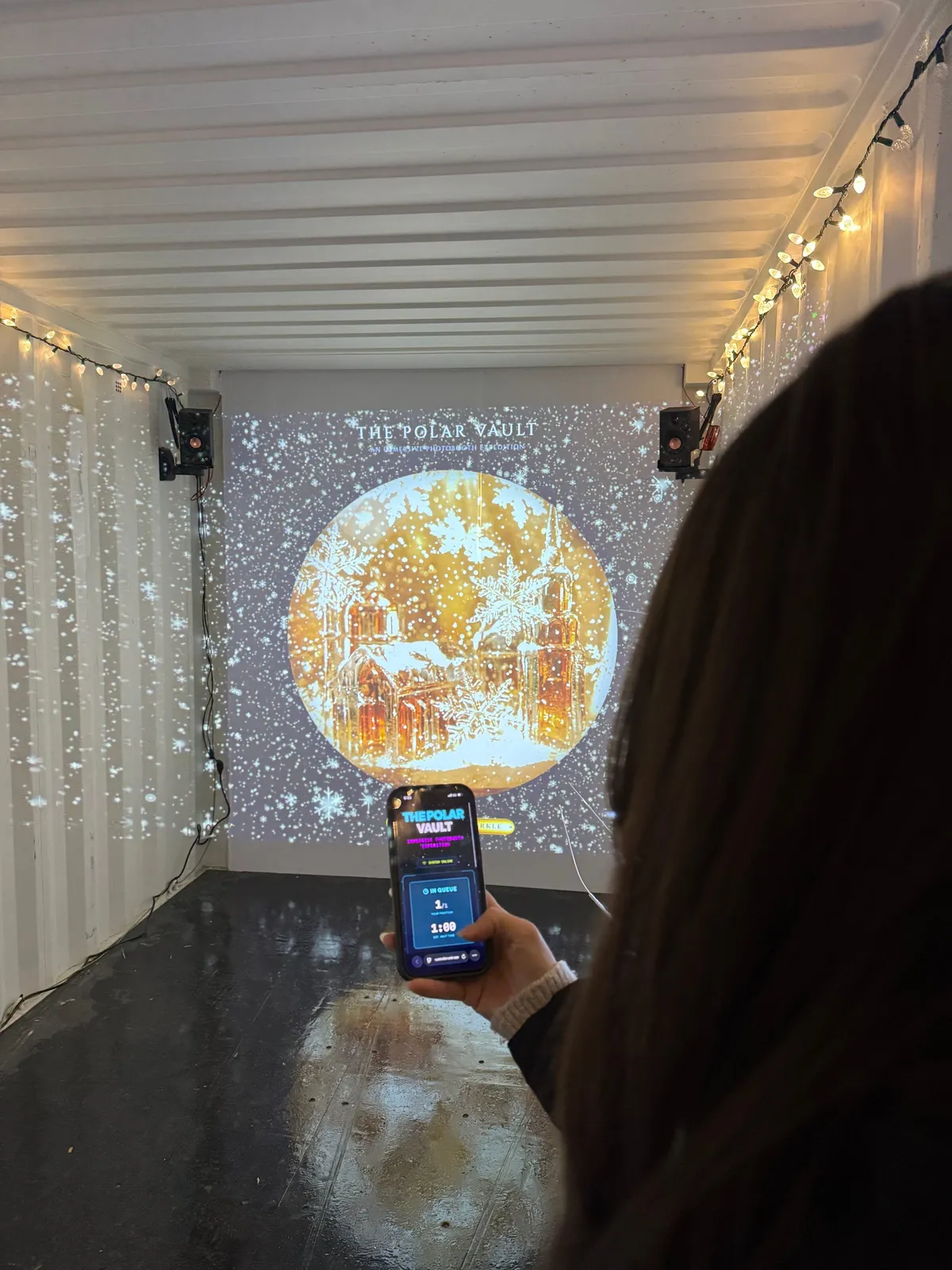 Inside the container, a visitor holds a phone showing the queue screen while a snow globe scene is projected on the wall.