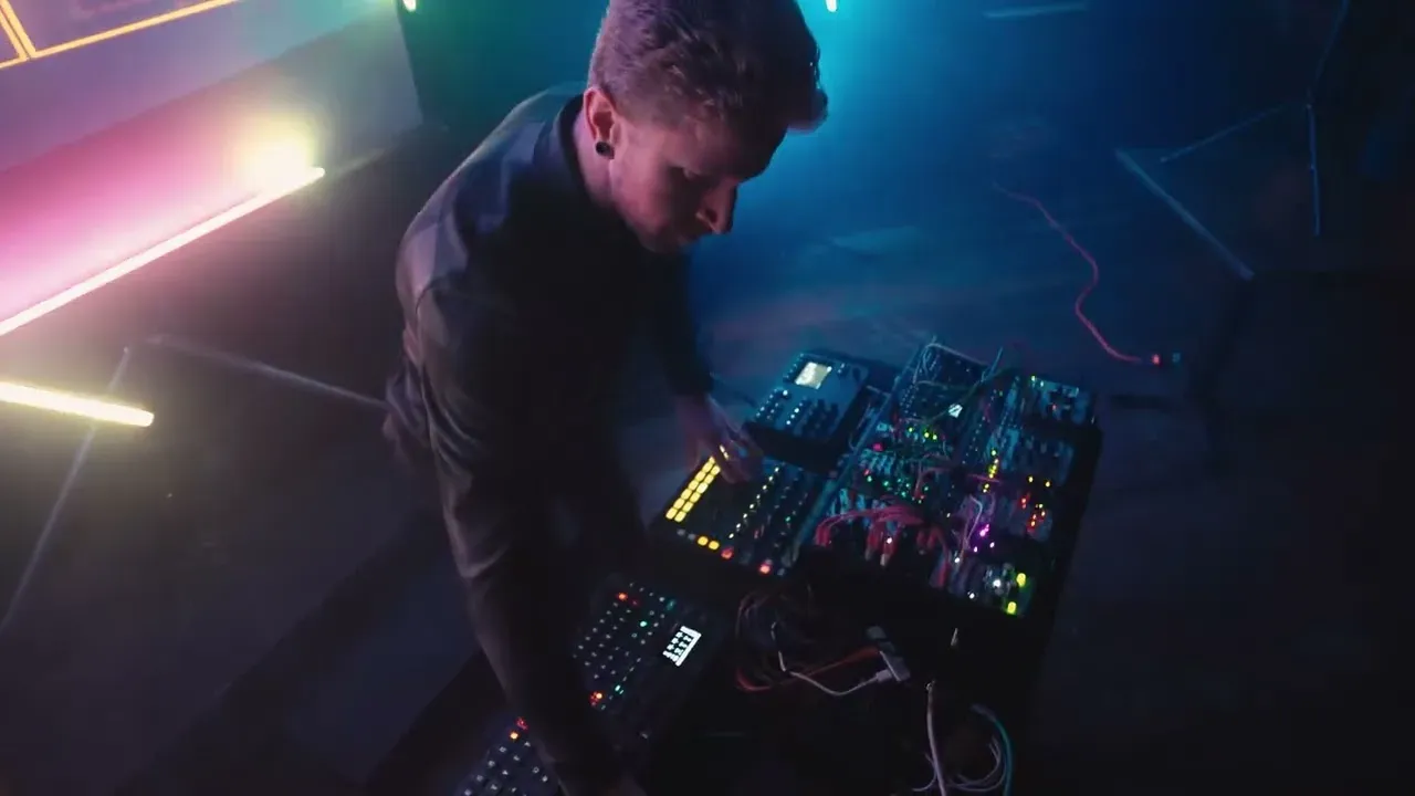 The performance rig seen from above — a modular synthesizer and controllers under coloured stage light, with two of the LED bars glowing pink and amber along the wall.